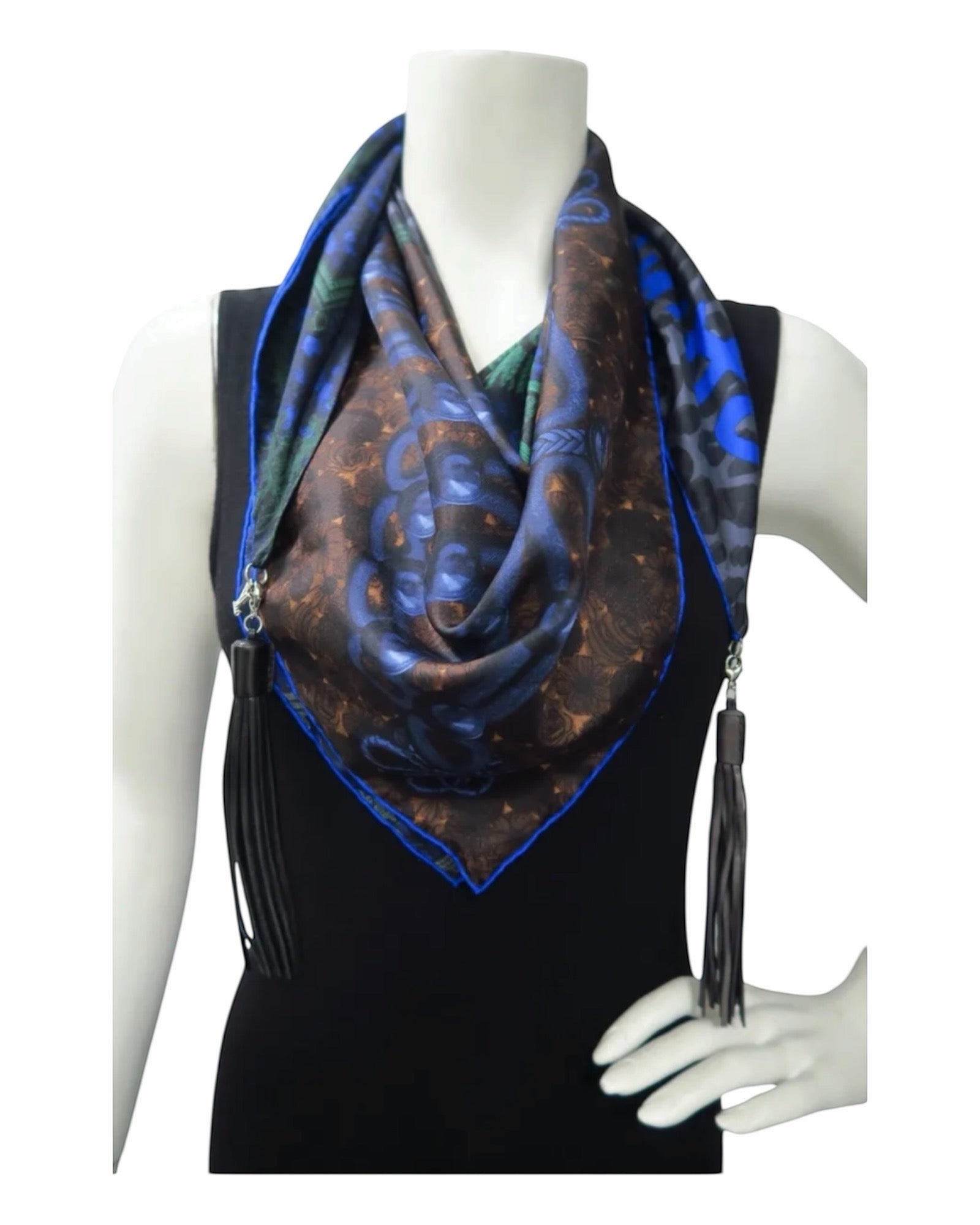 A square scarf with a blue and brown abstract pattern and black tassels, draped on a black sleeveless top displayed on a white mannequin torso.