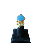 A gold ring with a large light blue hexagonal stone and smaller matching blue stones, displayed on a black velvet ring stand against a white background.